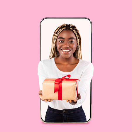 Smiling millennial black female gives box with gift from empty screen of huge phone, isolated on...