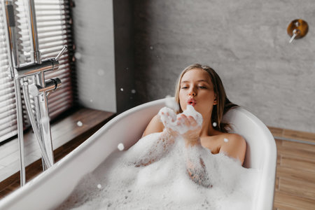 Young woman blowing soap bubbles and having fun while lying in bathtub full of foam at home, copy...