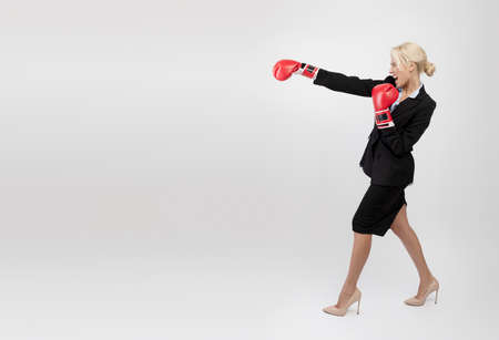 Angry young businesswoman in boxing gloves making strike, standing against light grey studio background with free spaceの写真素材