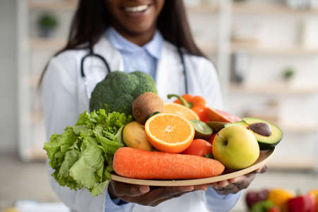 Black dietitian in lab coat holding bowl of fresh fruits and vegetables, recommending healthy plant based dietの写真素材