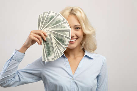 Portrait of happy business lady hiding face behind cash, holding bunch of dollars and smiling over...
