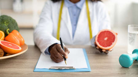 African american female dietologist holding grapefruit and writing healthy eating plan or weight loss diet for patientの写真素材