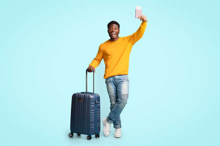 Cheerful african american guy with suitcase showing flight ticketsの写真素材