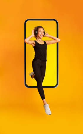 Sporty Lady Jumping From Phone Screen Wearing Headphones, Yellow Background