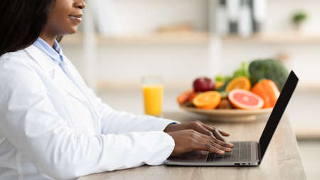 Black female dietitian with laptop consulting patient online at clinic, sitting at desk, side view, croppedの写真素材