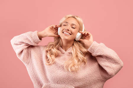 Peaceful young woman listening to music with closed eyes, wearing and touching headphones on pink studio backgroundの写真素材