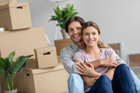 Smiling young caucasian husband hugging wife, rest, sitting in room interior with cardboard boxes with belongingsの写真素材