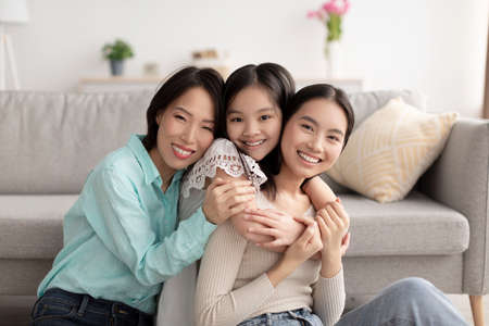 Young Asian woman with her cute daughter and mature mother hugging and smiling at camera indoors