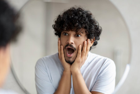 Skin problems concept. Shocked indian man looking at mirror and touching face, guy noticed acne, standing in bathroomの写真素材