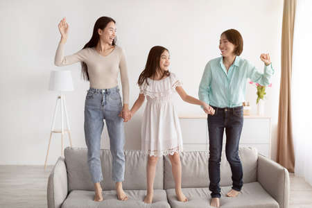 Happy little Asian girl with her mother and granny jumping on couch, having fun in living room