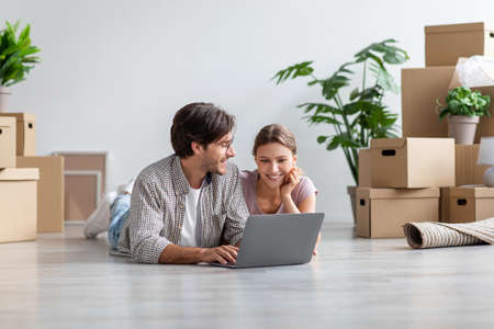 Pleased european millennial couple lie on floor look at computer among cardboard boxes with stuff in...