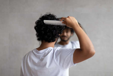 Back view of indian curly man combing hair standing near mirror in bathroom. Male haircare routine conceptの写真素材