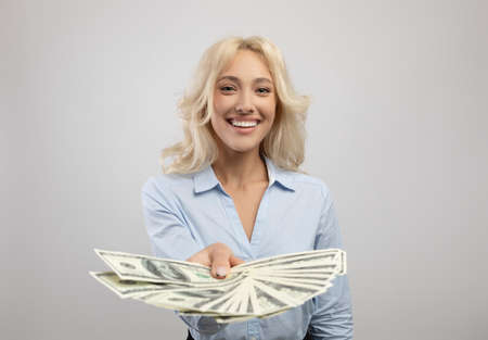 Happy young business lady holding fan of American money, showing banknotes to camera over light grey...