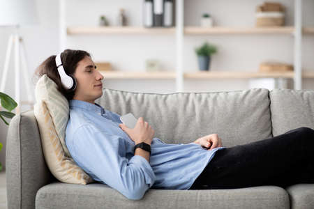 Rest and relax concept. Calm guy lying on sofa, listening to music, enjoying meditation for sleep in wireless headphonesの写真素材