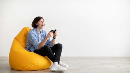 Young guy with joystick playing online video game, sitting in beanbag chair over light wall, panorama with free spaceの写真素材