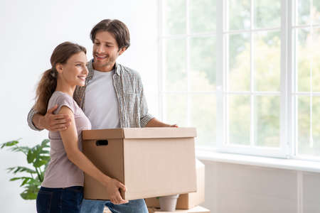 Smiling young european husband hug wife hold cardboard box with stuff in empty room interior