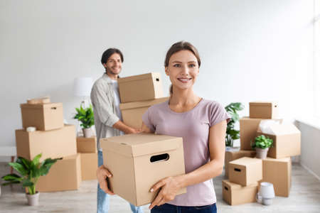 Smiling millennial caucasian lady and guy in casual clothes carry cardboard boxes with belongings in...