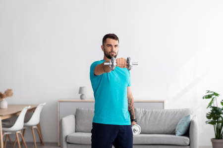 Serious millennial european man athlete in blue t-shirt raises dumbbell in minimalist living room...
