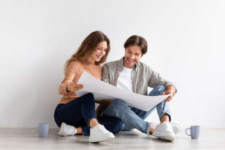 Cheerful young european man and lady sitting on floor and looking at house plan, planning repairs and movingの写真素材