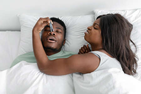 Dissatisfied funny millennial black lady wake up from snoring sleeping man, closes his nose with...