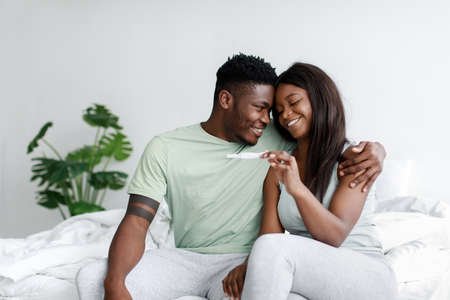 Glad happy young african american lady and guy look at pregnancy test on white bed in bedroom...