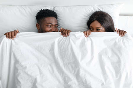 Happy funny young african american husband and wife covered with blanket and looking at each other...