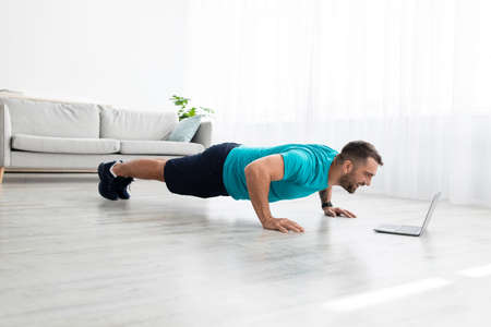 Cheerful millennial muscular european man doing push ups and watching online lesson on computer in...