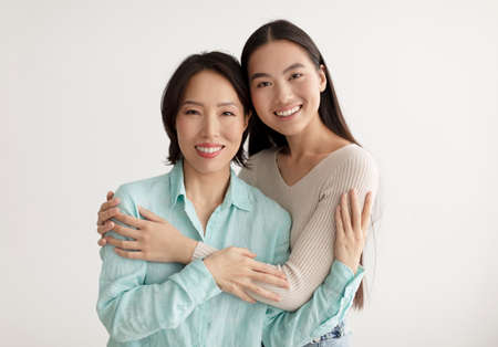 Joyful young Asian woman and her senior mother hugging and smiling at camera on white studio backgroundの写真素材