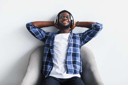 Relaxed African American guy listening to music in wireless headphones, resting on armchair with...