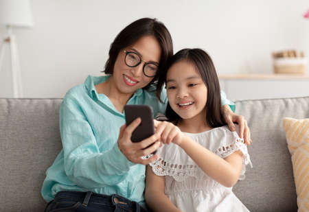 Loving Asian grandmother and her cute little granddaughter using smartphone on couch at homeの写真素材