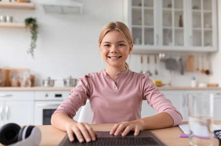 Portrait of smiling caucasian adolescent girl blonde typing on keyboard by laptop in kitchen...