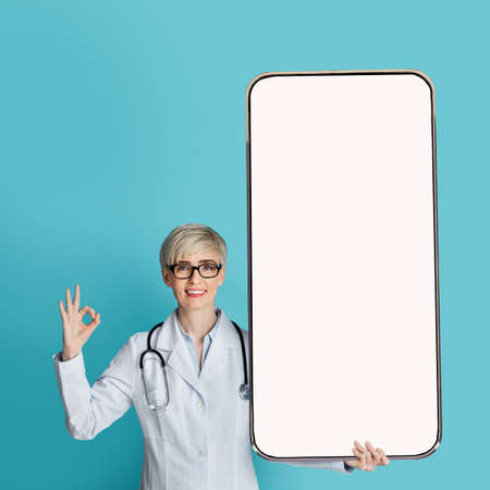 Female doctor showing big smartphone with blank screen and gesturing OK sign, standing over blue background, mockupの写真素材