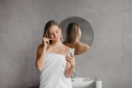 Pretty lady wrapped in towel using cellphone while doing morning routine in bathroom, brushing teethの写真素材