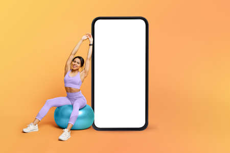 Fitness app. Athletic black lady training on fitness ball near big cellphone with blank screen,...