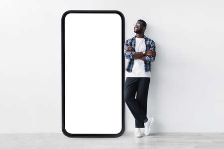 Full body length of black guy standing near huge giant smartphone with empty white screen template, mockupの写真素材