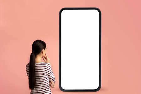 Back view of woman looking at big smartphone with blank white screen and thinking, device with space...