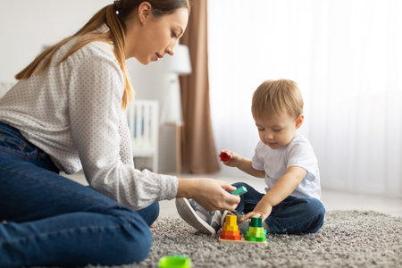Young mom and toddler child playing together at home, using toys while sitting on floor carpet in living roomの写真素材