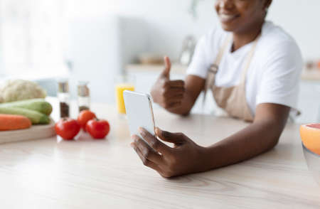 Smiling young black lady show thumb up, take photo on phone on table with vegetables in kitchen interiorの写真素材