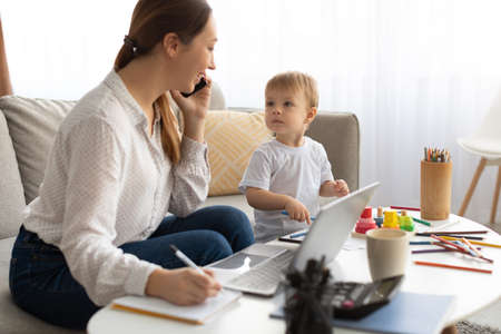 Young busy mother working from home with kid, talking on cellphone and making notes in notepadの写真素材
