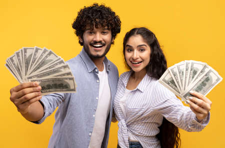 Big luck and win concept. Excited young indian couple holding dollar cash, posing with money on yellow backgroundの写真素材