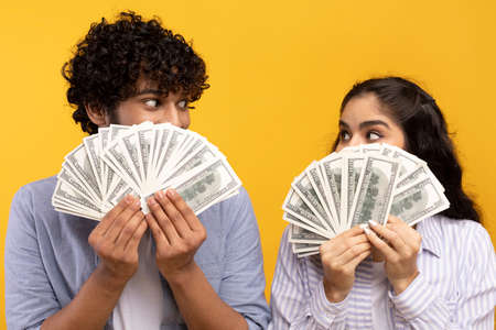 Big win concept. Young indian couple holding and covering faces with a lot of dollar cash fan,...