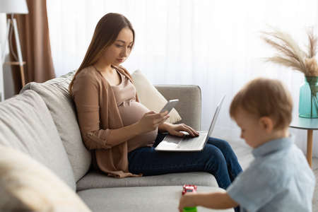 Young pregnant freelancer woman using smartphone and laptop, mom taking care of her toddler sonの写真素材