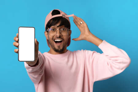 Emotional hindu guy in baseball cap showing cell phone, mockup