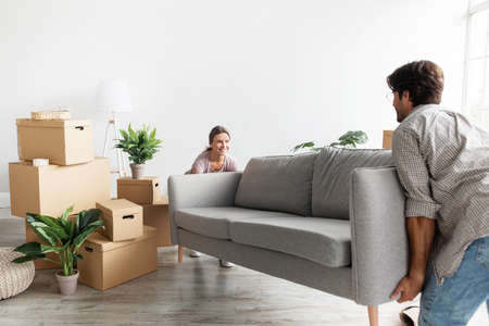 Smiling millennial caucasian couple in casual carry sofa in room with packed cardboard boxes,...