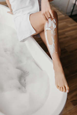Young woman shaving legs using safety razor, removing hair, making depilation in bathroom, sitting...