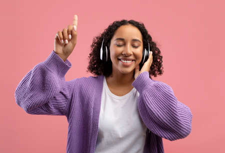 Beautiful young black woman listening to music in wireless headphones, closing eyes and pointing...