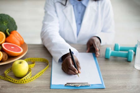 Cropped view of black female dietitian planning meal for client, writing in clipboard, sitting at...