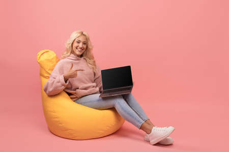 Cool website or ad. Happy woman pointing at laptop with blank screen, sitting on beanbag chair, pink background, mockupの写真素材