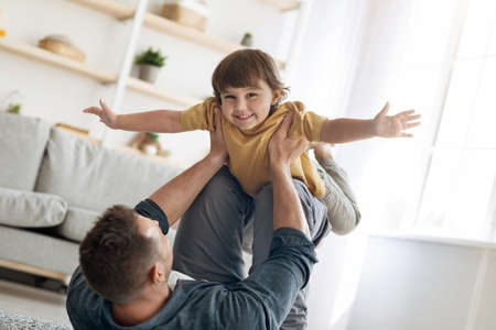 Fun with daddy. Portrait of adorable little boy playing with father at home, man lifting kid up on legs at homeの写真素材