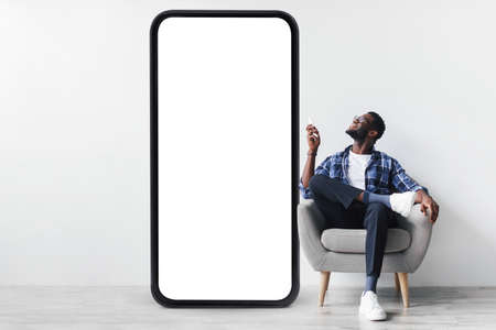 Smart home system. Young black guy with remote control sitting in armchair near huge smartphone, mockup for designの写真素材